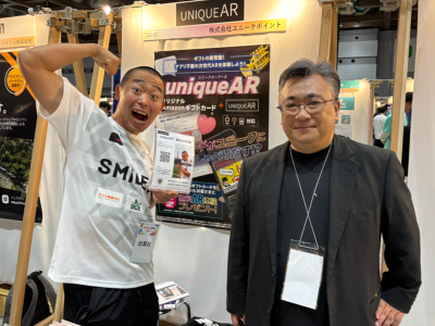ユニークポイント 代表取締役 薬師丸勇人氏（右）。同氏はサッカー芸人オールスターチーム「スマイラーズ」のスポンサーを務める関係で展示会にはシイナの三浦 一馬さんが助っ人した。三浦さんは春日さんが結婚するまで約20年間暮らしていた「むつみ荘」で生活しているという